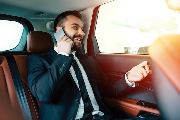 Улыбающийся бизнесмен в полном костюме разговаривает по телефону, сидя на заднем сиденье машины такси