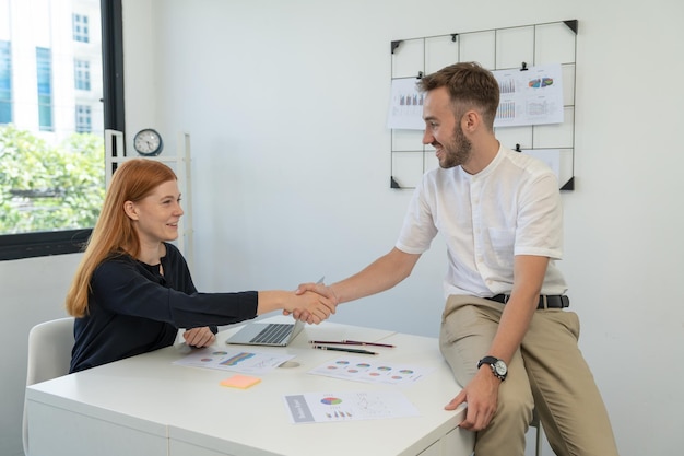 Smiling businessman handshake young businesswoman colleague after discussing agreement for marketing planning strategy on her desk in office