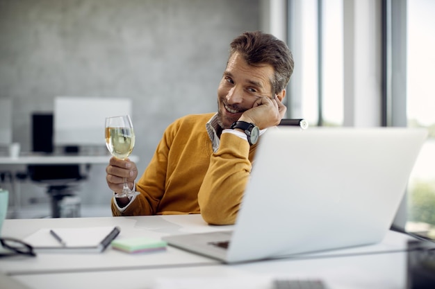 Smiling businessman having glass of wine while talking during\
video call in the office