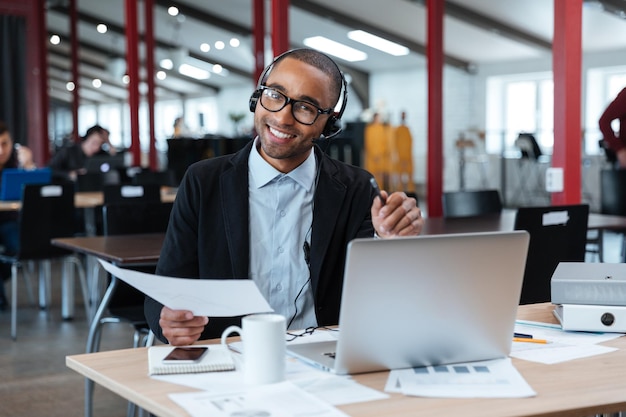 Smiling businessman in headphones holding documents and pointing at you at the office