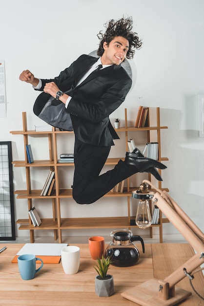 Photo smiling businessman jumping near workplace with coffee pot and tablet in office