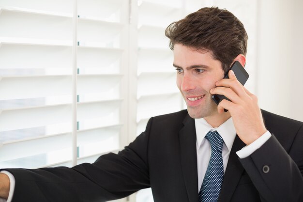 Smiling businessman peeking through blinds while on call