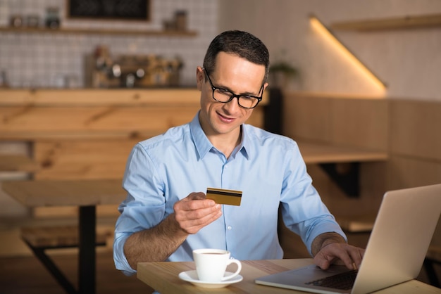 Улыбающийся бизнесмен делает покупки в Интернете с помощью кредитной карты с помощью ноутбука в кафе