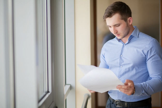 Smiling businessman standing by the window in office and reading paper