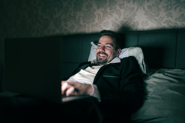 밤에 호텔 방에 침대에 누워 작업을 위해 노트북을 사용하는 소송에서 사업가 웃고. 과로 개념.