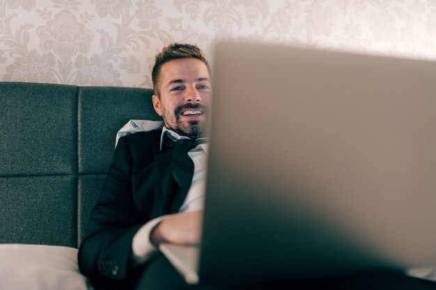 Улыбающийся бизнесмен в костюме, лежа на кровати в гостиничном номере ночью и используя ноутбук для работы. Концепция переутомления.