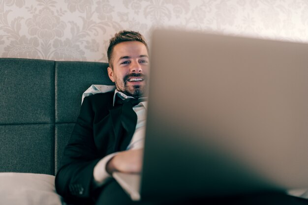 Smiling businessman in suit lying on the bed in hotel room at night and using laptop for work. Overworking concept.