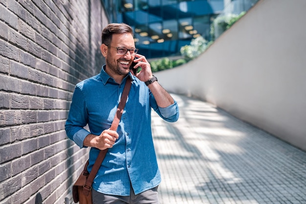 Smiling businessman talking on smart phone while walking by wall on footpath