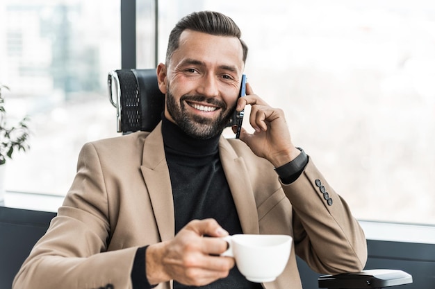 携帯電話で話し、カメラを見てコーヒーマグカップで笑顔のビジネスマン