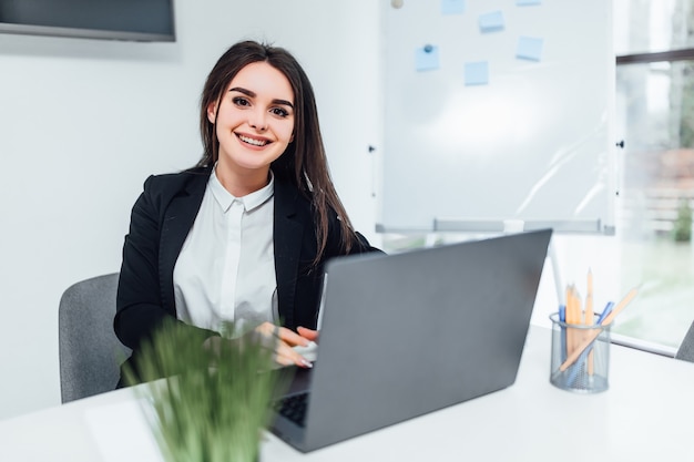 Smiling businesswoman dressed casual  suit sitting in modern office and using laptop.