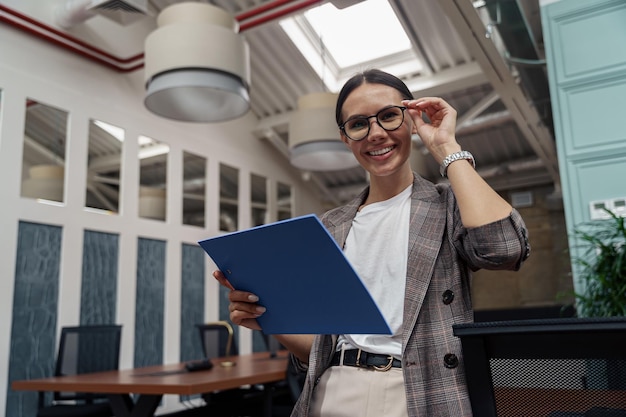 Улыбающаяся деловая женщина в очках с планшетом стоит в офисе и смотрит в камеру
