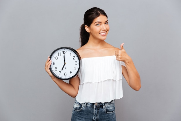 Smiling businesswoman holding clock and showing thumb up isolated on a gray wall