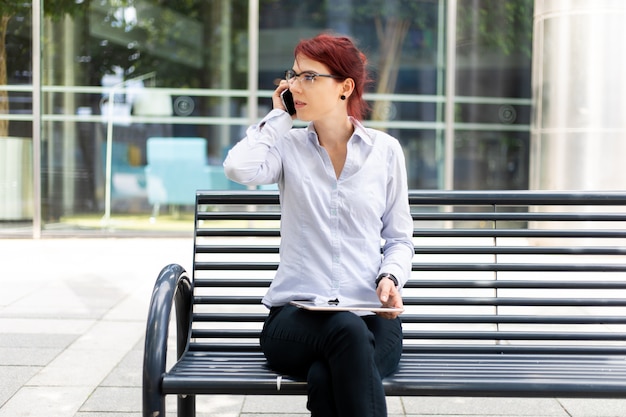 Smiling businesswoman using a digital tablet outdoor sitting on a bench and talking on the phone