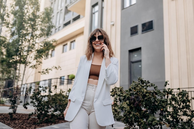 Smiling businesswoman in white suit talking phone during walking in city with modern architecture