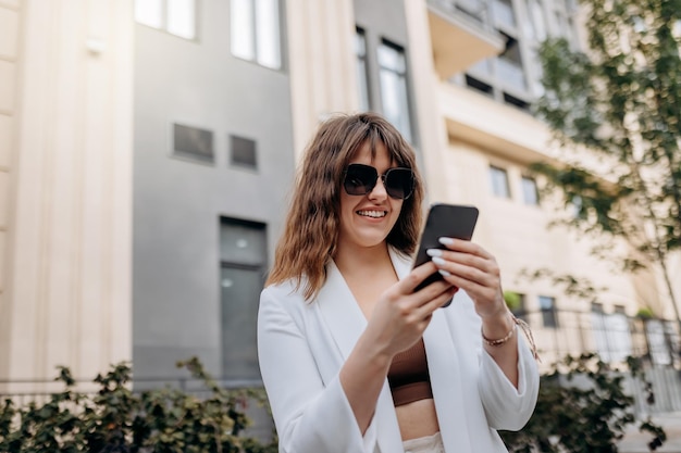 Smiling businesswoman in white suit using phone during walking in city with modern architecture