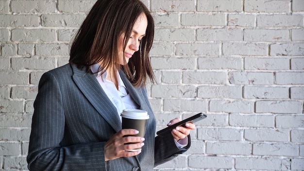 Photo smiling businesswoman with short dark hair types on black smartphone holding paper coffee cup standing near white brick wall in morning copy space