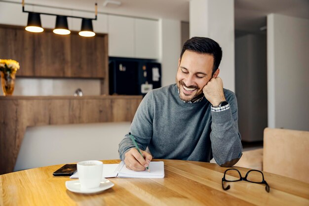 A smiling busy man is writing tasks in textbook at home