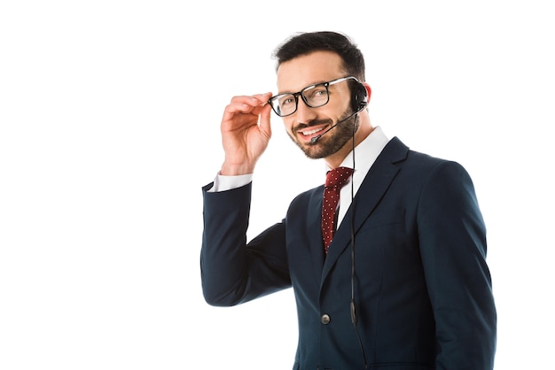 Smiling call center operator in headset looking at camera and touching glasses isolated on white