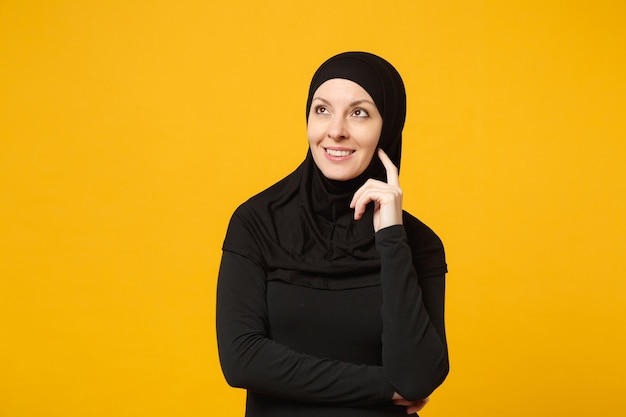 ヒジャーブの黒い服を着た穏やかな若いアラビアのイスラム教徒の女性の笑顔は、黄色の壁、肖像画で孤立して見上げて、手を組んで保持します。人々の宗教的なライフスタイルの概念。コピースペースのモックアップ