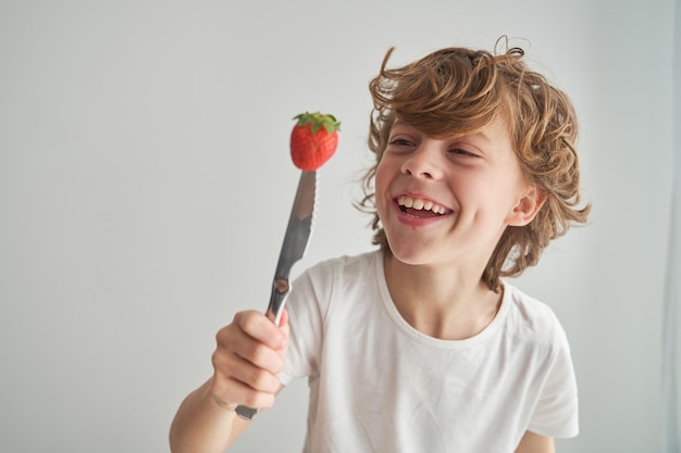 Smiling carefree child with curly hair having fun while looking at ripe berry pierced with knife on white background