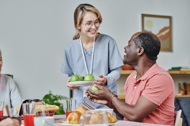 Smiling caregiver giving apples to senior man while he sitting at table and eating his breakfast