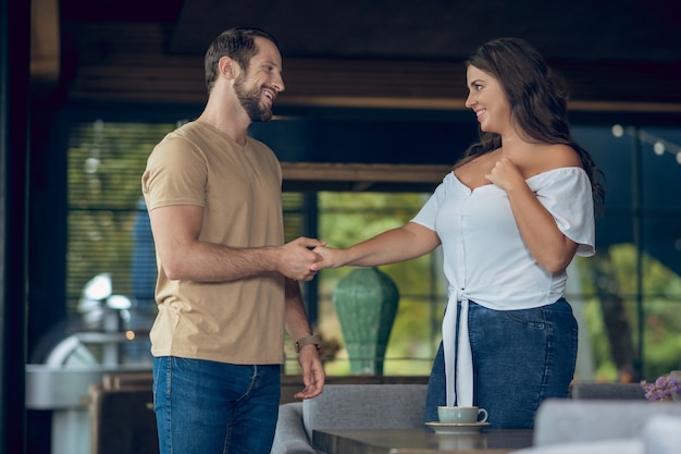 カフェのテーブルに座っている長い髪の楽しい女性の肩を抱き締める思いやりのある男の笑顔