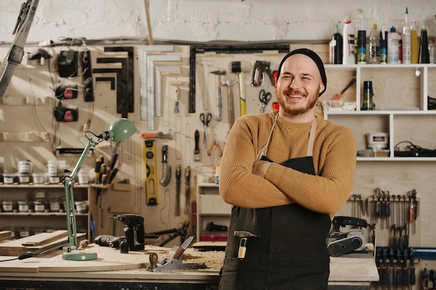 Smiling carpenter in a watch cap posing for a photo, standing\
with hands crossed in a big workshop number of tools hanging on the\
wall.