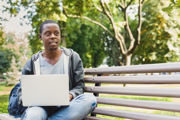 Smiling casual african-american student working with laptop in the park in pleasant atmosphere. Technology, communication, education and remote working concept, copy space