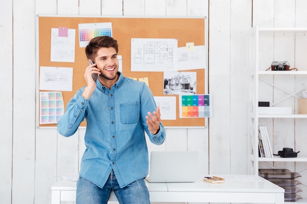 Smiling casual businessman sitting on the office desk and talking on mobile phone