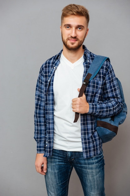 Smiling casual man with backpack looking at front isolated on a gray wall