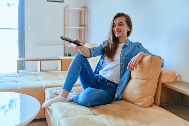 Smiling casual modern joyful happy young girl sitting on a sofa, holding remote and watching TV a t home