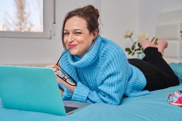 Smiling casual young woman using laptop in bed looking at camera