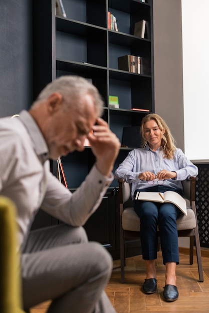 Smiling Caucasian blonde woman holding eyeglasses in her hands while sitting on a chair in front of a tired man