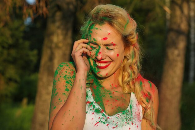 Smiling caucasian blonde woman with bright makeup posing covered by Holi paint