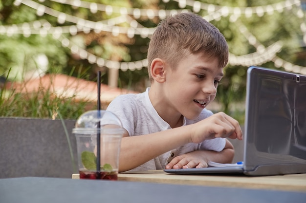 Smiling caucasian boy sitting at table at veranda of cafe studying with laptop