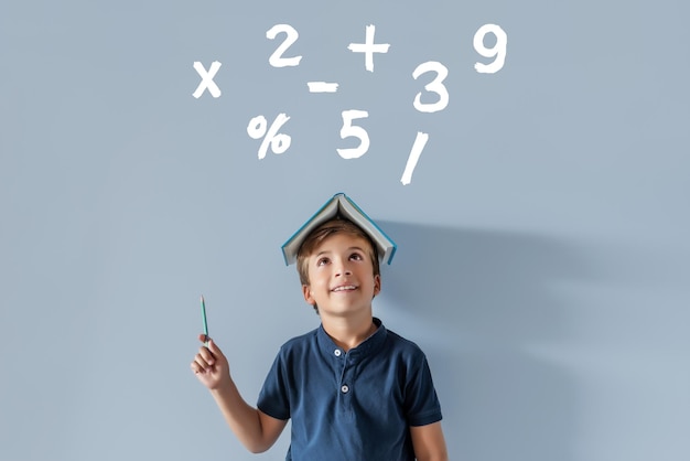 Фото Улыбающийся кавказский мальчик с книгой на голове, указывающей на математические символы снова в школу