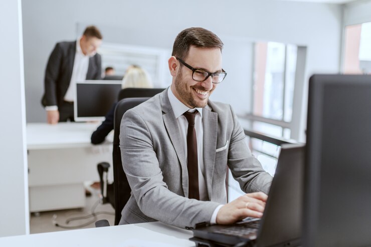 Photo smiling caucasian businessman in suit and with eyeglasses sitting at his workplace and using computer, hands on keyboard.