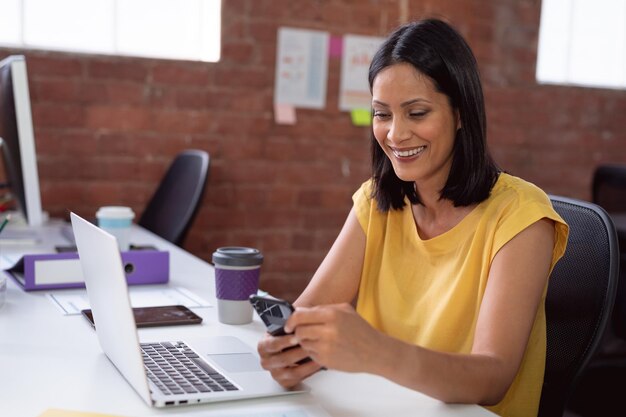 Smiling caucasian businesswoman sitting at desk in office with laptop, using smartphone