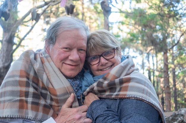 Smiling caucasian elderly couple lovingly hugs each other in the woods looking at camera