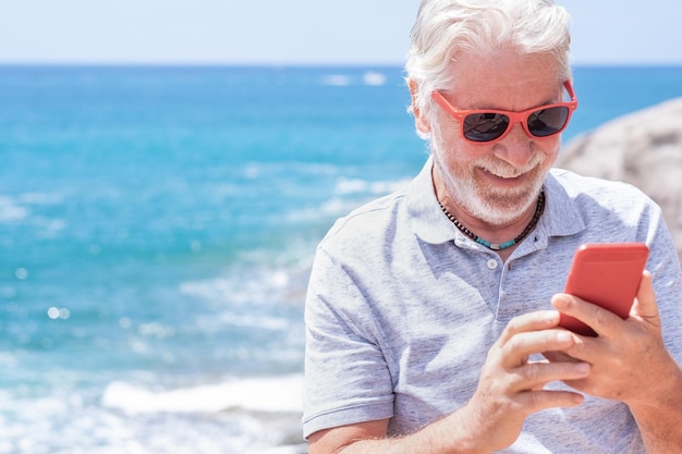 Smiling caucasian grandfather in outdoors at sea enjoying sunny\
day and vacation using mobile phone senior man with red sunglasses\
using modern technologies and wireless connection