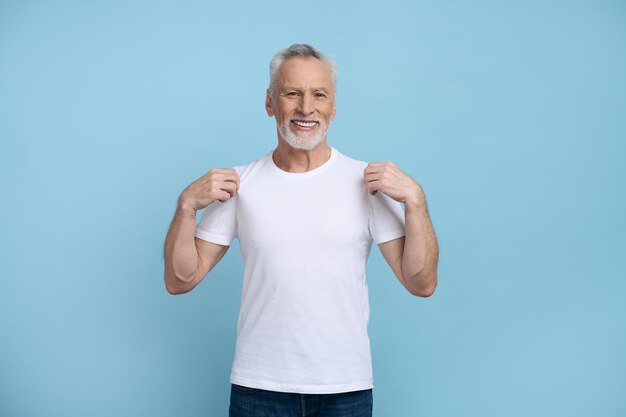 Smiling caucasian gray haired bearded senior man showing a white tshirt he's wearing with mockup copy space for your ads smiling looking at camera isolated over blue background