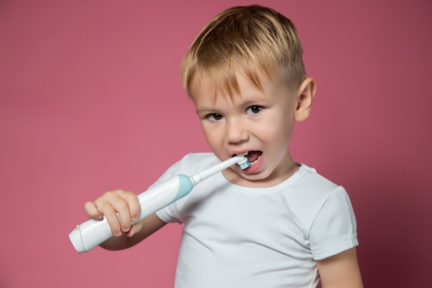 Smiling caucasian little boy cleaning his teeth with electric sonic toothbrush on pink background.