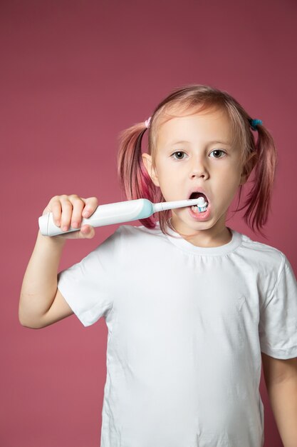 Bambina caucasica sorridente che si pulisce i denti con uno spazzolino sonico elettrico su sfondo rosa.