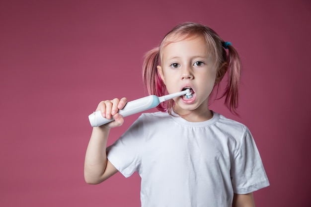 ピンクの背景に電気音波歯ブラシで彼の歯を掃除している白人の少女の笑顔。