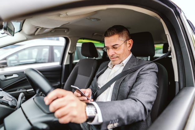 L'uomo caucasico sorridente ha vestito casuale astuto con la cintura di sicurezza sopra e con la mano sul volante che fa causa allo smart phone per la scrittura o la lettura del messaggio. interni auto.