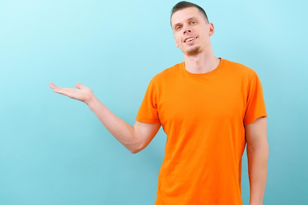 A smiling caucasian man in an orange tshirt pointed a hand on the middle side Showing empty copy space on blue background Handsome Joyful Model Present Product Confidence Joy Copy Empty