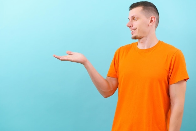 A smiling caucasian man in an orange tshirt pointing and looking at a hand on the middle side Showing empty copy space on blue background Portrait Young Adult Hand Smile Arm