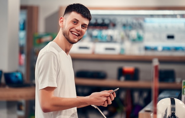 Smiling caucasian man in white t-shirt trying out new smart phone. Tech store interior.