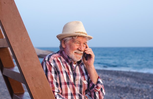 Smiling caucasian senior man on the beach looking at the golden hour of sunset at sea while talking on mobile phone bearded male enjoying free time vacation or retirement