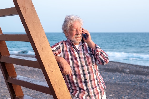 Smiling caucasian senior man on the beach looking at the sunset light at sea while talking on phone